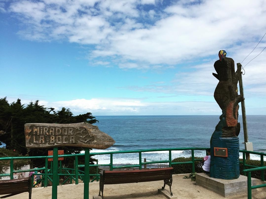 Fotografía del mirador, se aprecia un letrero con su nombre, una estatua y el borde del mar, desde un sector más alejado